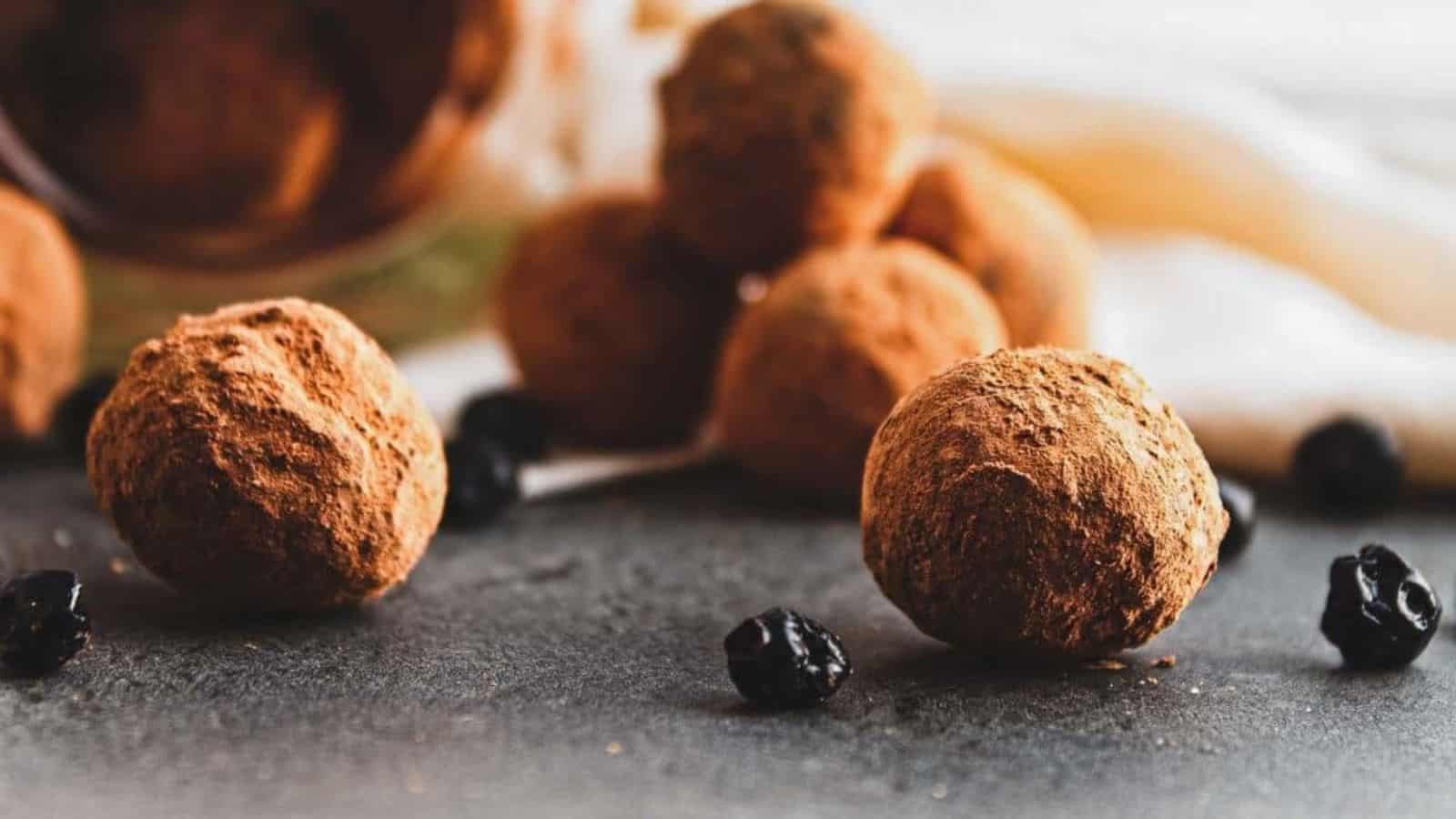 Chocolate truffles coated with cocoa powder are placed on a gray surface. Some of the truffles and small dried berries are in the background, slightly out of focus. A light fabric is draped behind them.