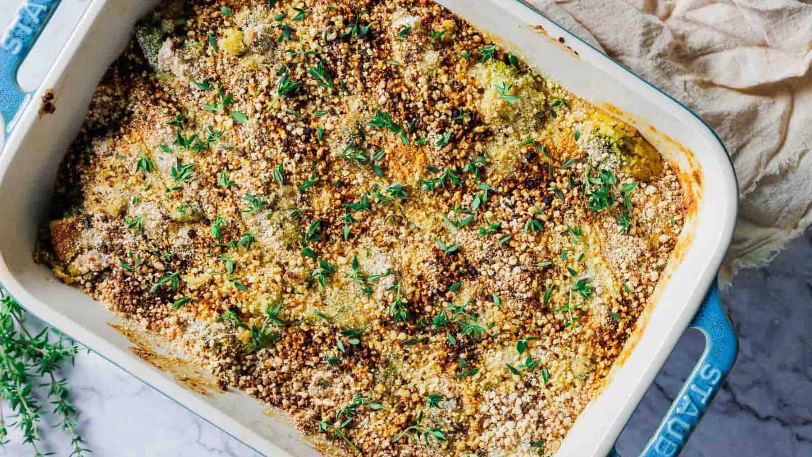 A black plate with a serving of broccoli casserole, topped with breadcrumbs. A fork rests on the plate. The background is a marble surface, with some green plant decorations partially visible.