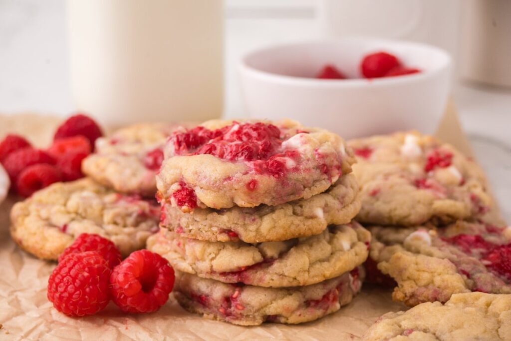 A stack of raspberry cookies with white chocolate chips on parchment paper, surrounded by fresh raspberries and a bowl of raspberries in the background.