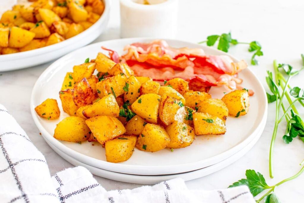A white plate with cooked diced potatoes garnished with herbs and several strips of crispy bacon, with fresh parsley and another plate of potatoes in the background.