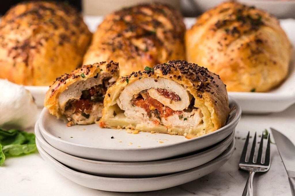 A sliced stuffed chicken breast wrapped in golden pastry is served on stacked plates, with more whole pastries in the background and utensils beside the plate.