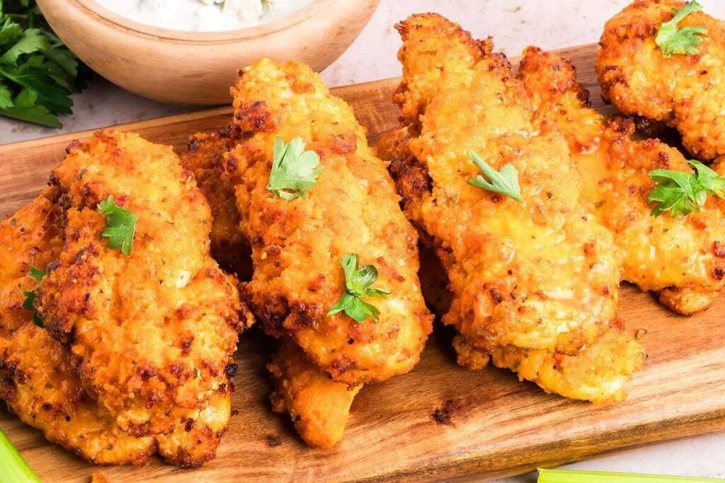 A wooden board with several crispy fried chicken tenders, garnished with parsley, with a bowl of dipping sauce and celery sticks in the background.