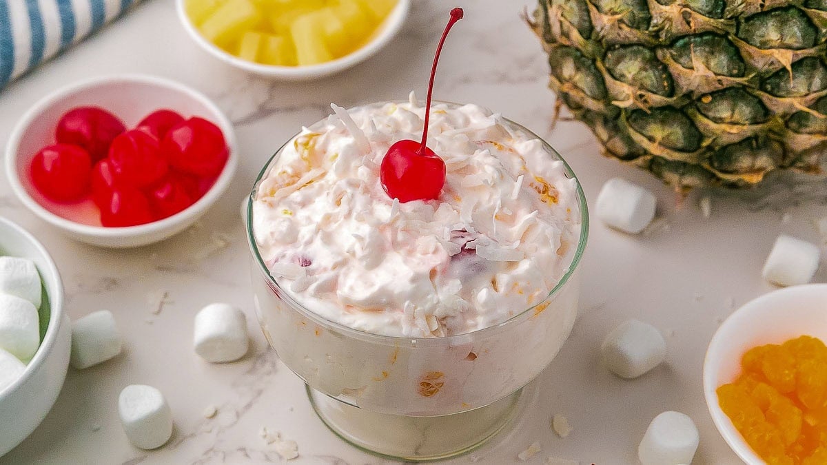 A glass bowl of ambrosia salad topped with a cherry, surrounded by marshmallows, pineapple, cherries, and a whole pineapple on a marble surface.