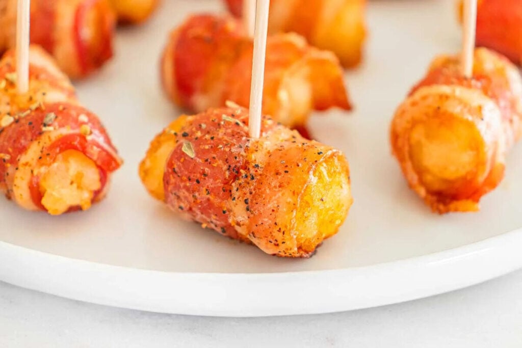 Bacon-wrapped tater tots secured with toothpicks are arranged on a white plate, seasoned with black pepper and herbs—perfect as tasty appetizers for any gathering.