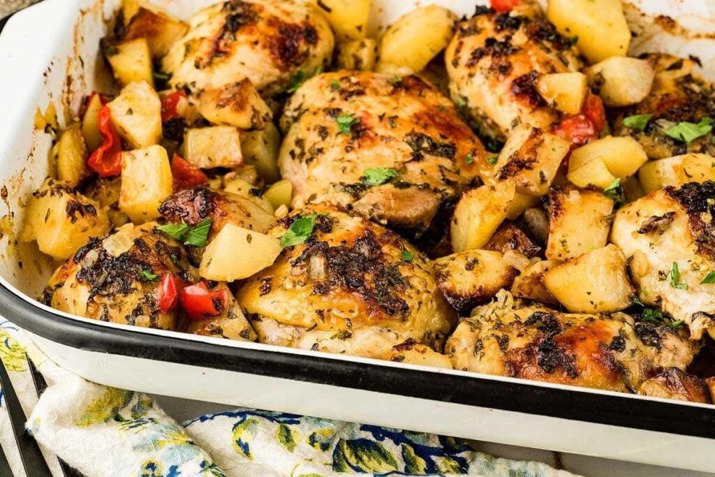 Baked chicken thighs with browned skin, chunks of potatoes, and pieces of red bell pepper in a white roasting pan, garnished with herbs.
