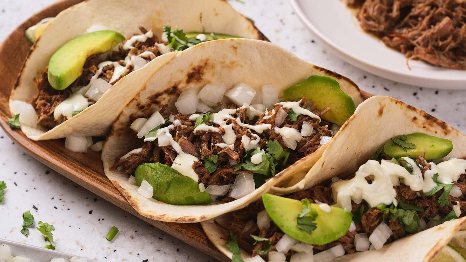 Three barbacoa tacos garnished with avocado, cilantro and white chopped onions next to a plate of shredded beef on the background.