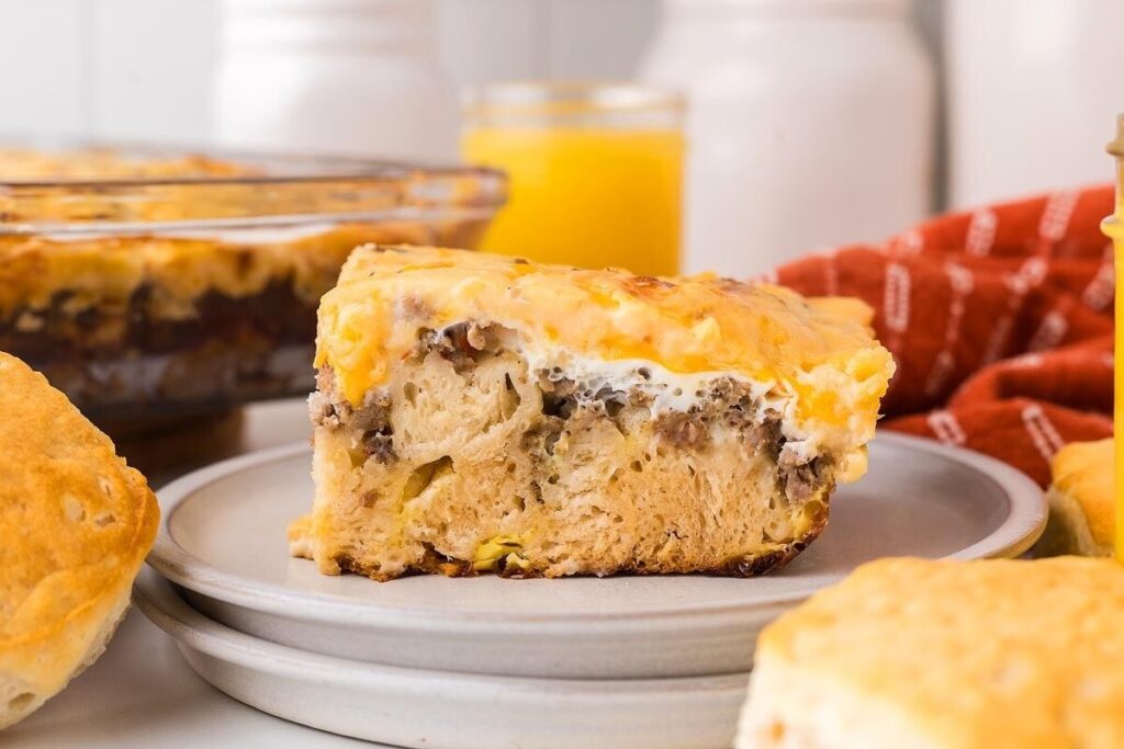 A slice of breakfast casserole with sausage, eggs, and cheese sits on a plate with biscuits and orange juice nearby.