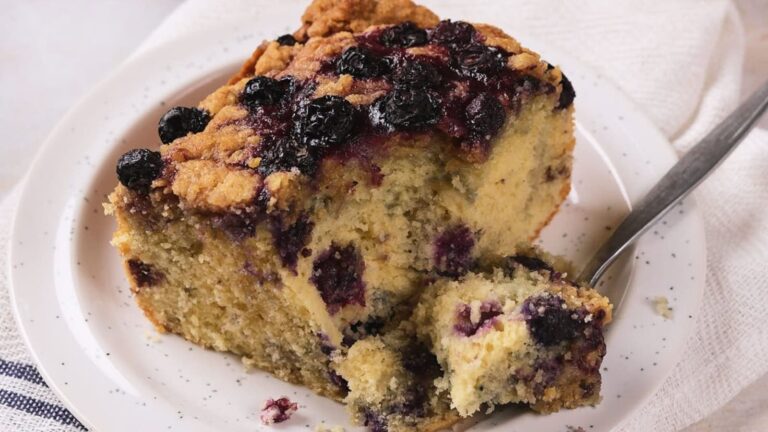 A slice of blueberry coffee cake on a white plate with a fork, showing blueberries and a crumbly texture.
