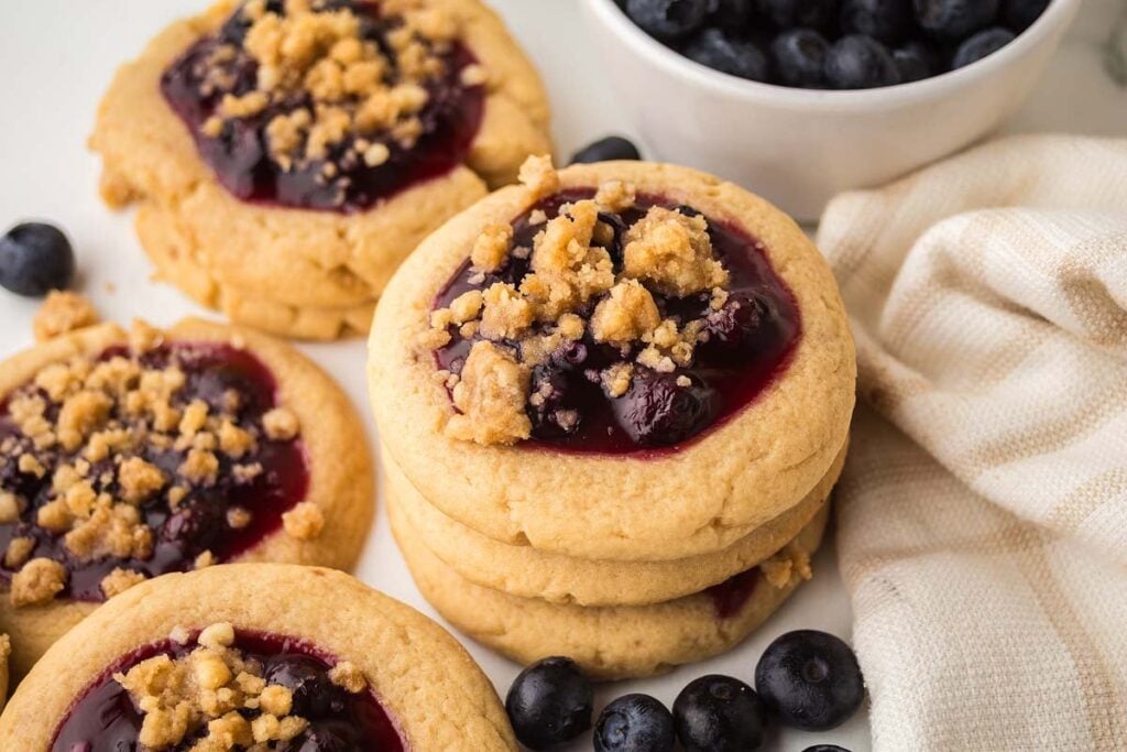 Three stacked cookies with blueberry jam and crumb topping, surrounded by more cookies, fresh blueberries, and a bowl of blueberries next to a beige cloth.