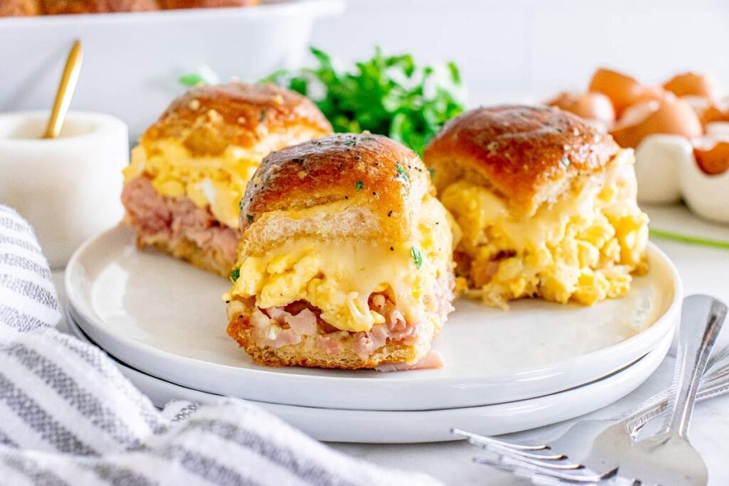 Three breakfast sliders filled with scrambled eggs, ham, and melted cheese are arranged on a white plate, with a garnish and eggs visible in the background.