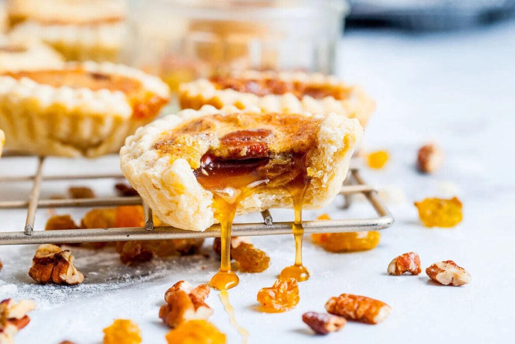 A butter tart with caramel filling oozing out sits on a cooling rack, surrounded by pecans and raisins on a light surface.