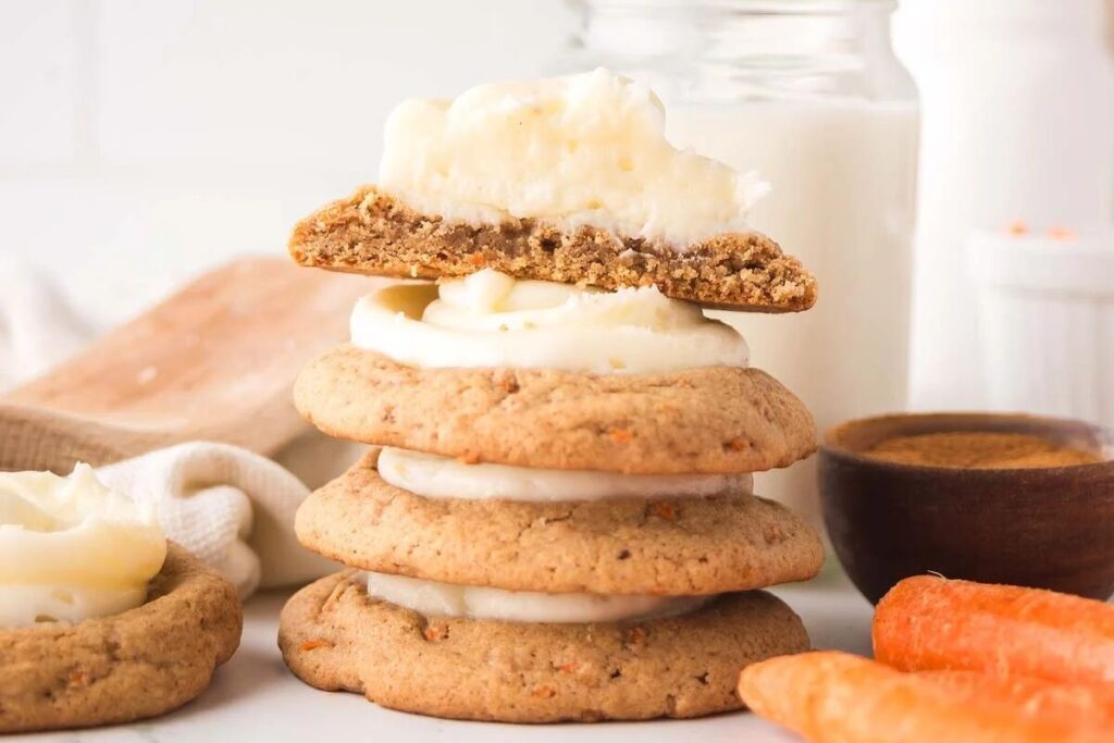 A stack of carrot cake cookies with cream cheese frosting, a halved cookie on top, surrounded by carrots, a wooden bowl of spice, and a glass of milk in the background.