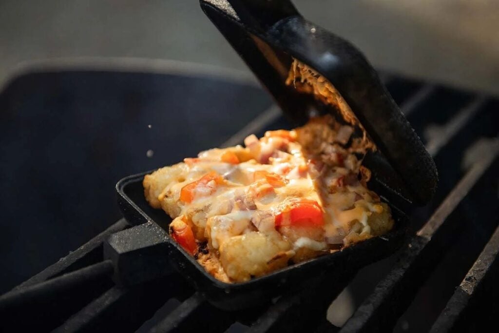 A close-up of a cast iron pie iron on a grill, filled with melted cheese, diced tomatoes, onions, and tater tots.