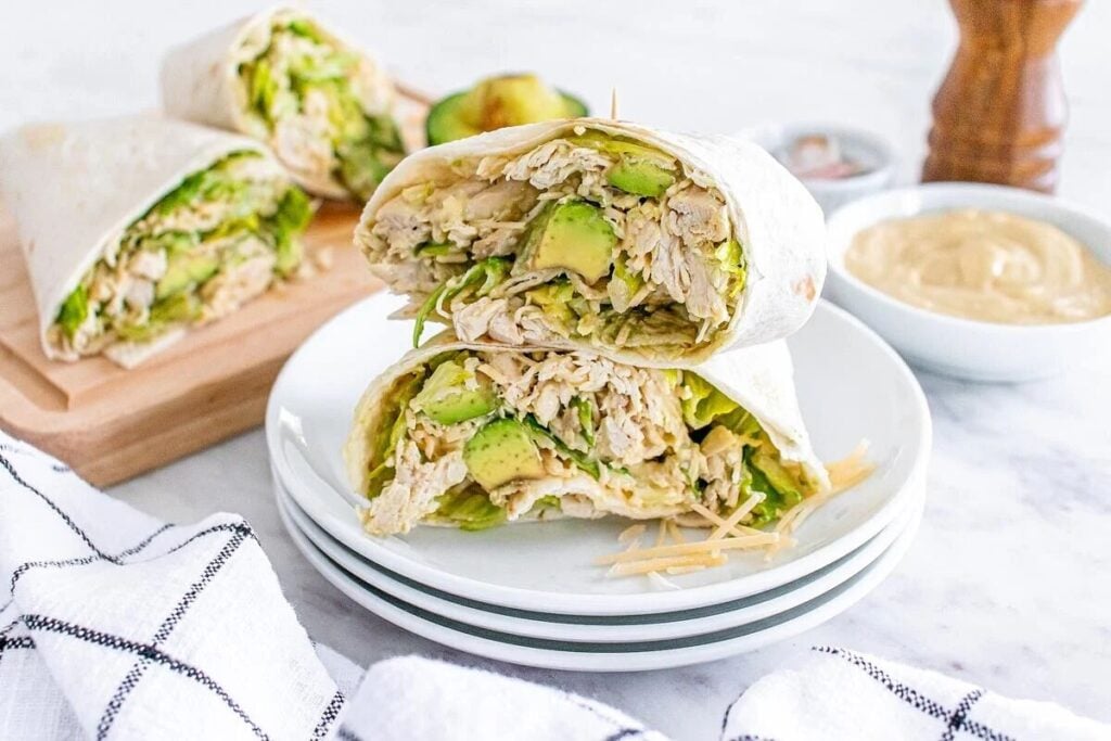 Two halved chicken avocado wraps stacked on a white plate, with sauce, a pepper grinder, and additional wraps in the background.