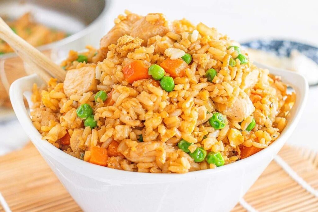 A white bowl filled with fried rice, mixed with peas, carrots, and chicken pieces, with a wooden spoon resting inside.