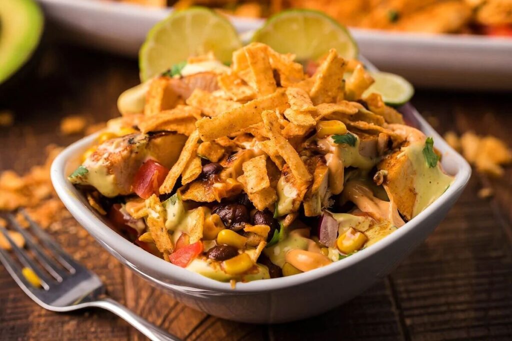 A bowl filled with grilled chicken, black beans, corn, diced tomatoes, sauce, and topped with crispy tortilla strips, with lime slices in the background.