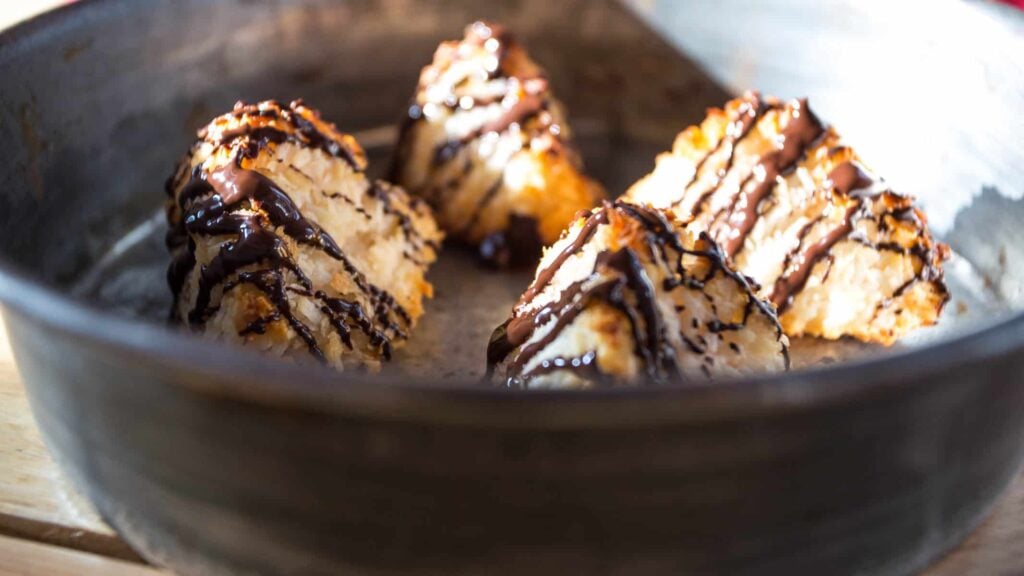 Four coconut macaroons drizzled with chocolate sauce are arranged in a round metal baking dish.