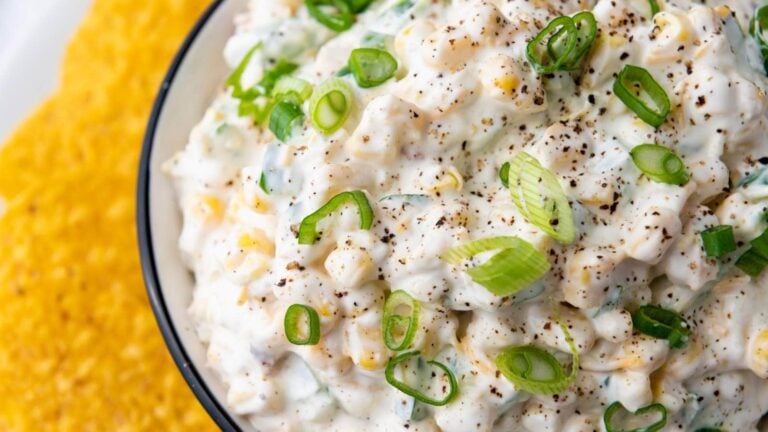 Close-up of a bowl of creamy corn salad garnished with sliced green onions and black pepper.