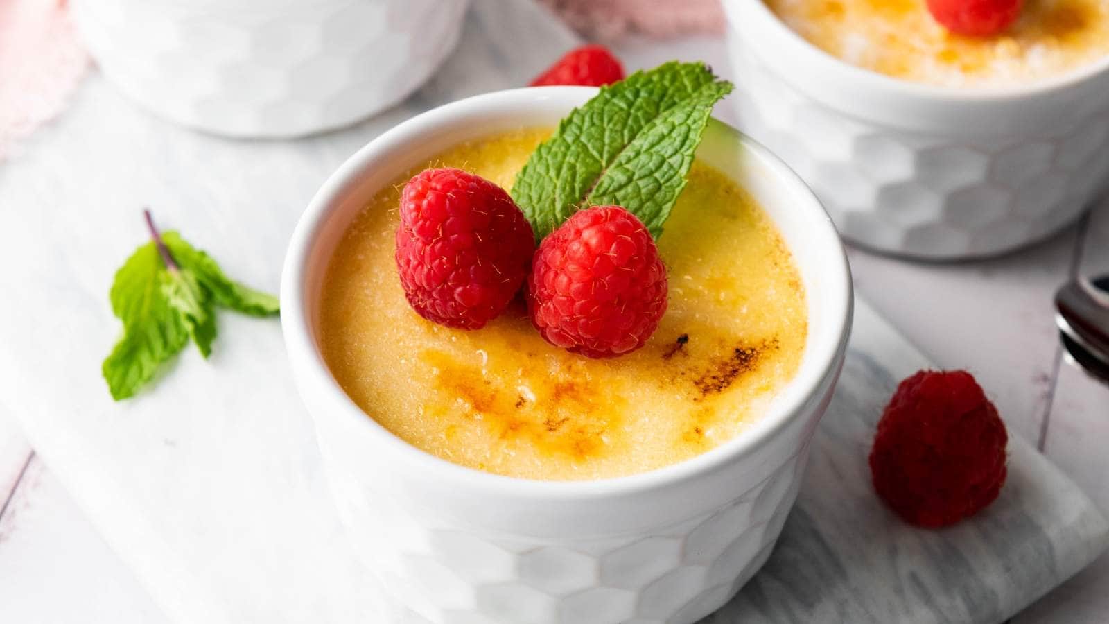 A ramekin of crème brûlée topped with two fresh raspberries and a mint leaf.