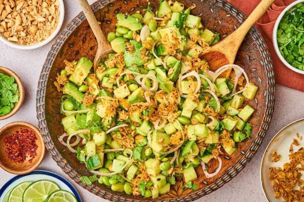 A bowl of chopped fresh vegetables and herbs, including cucumbers, avocado, edamame, and shallots, with two wooden utensils and small bowls of toppings on the side.