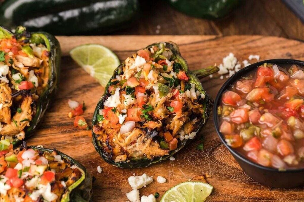 Stuffed poblano peppers filled with shredded chicken, topped with pico de gallo and cheese, served with lime wedges and a bowl of salsa on a wooden board.