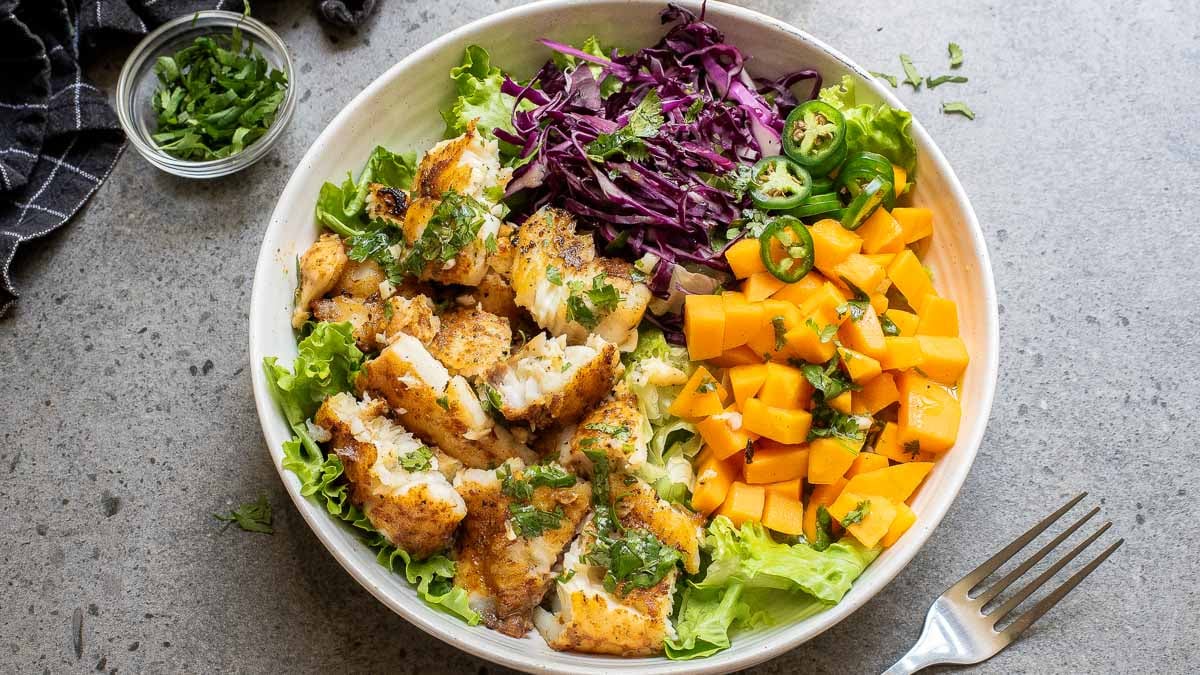 A bowl with grilled fish, lettuce, diced mango, red cabbage, jalapeño slices, and cilantro.