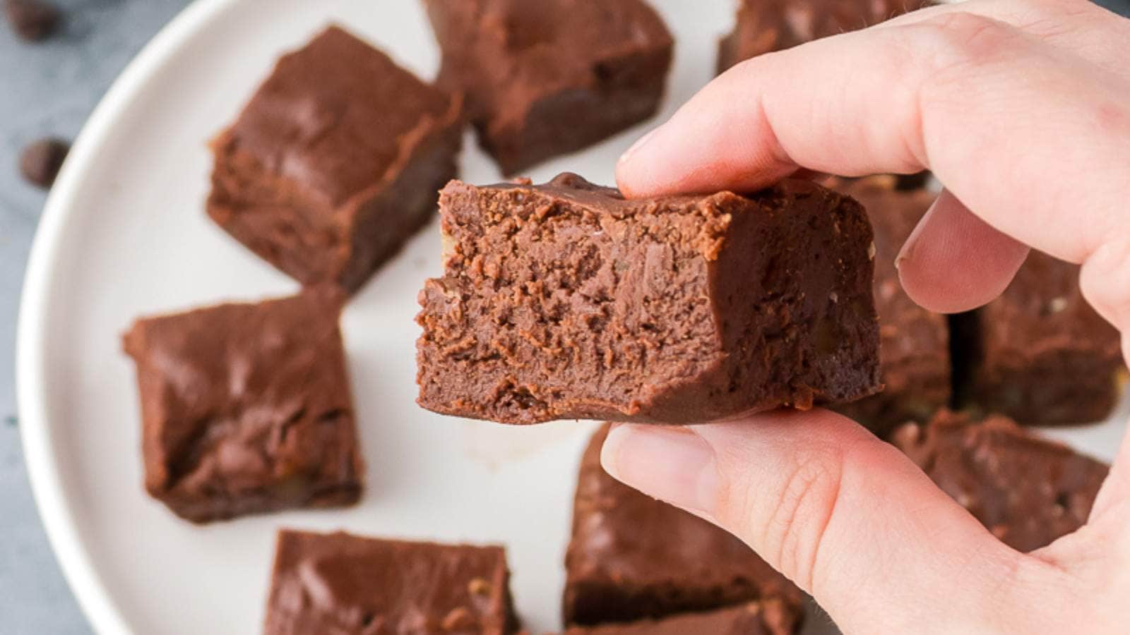 A hand holds a piece of chocolate fudge with a bite taken out, with more pieces of fudge on a white plate in the background.
