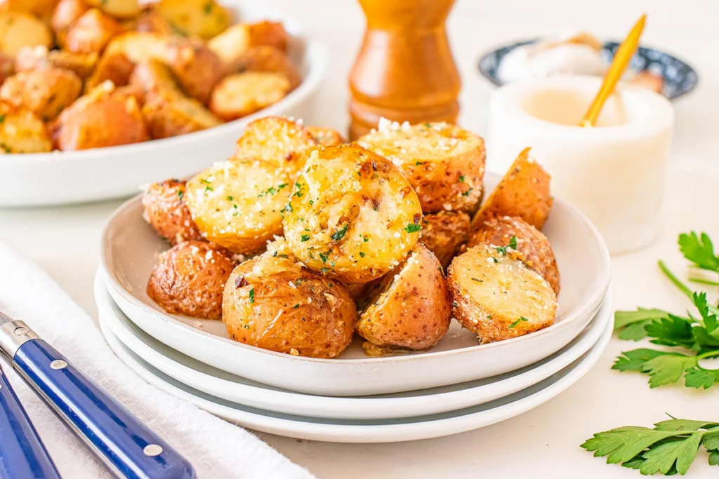 A plate of roasted baby potatoes garnished with herbs and grated cheese sits on stacked white dishes, with utensils and seasonings in the background.