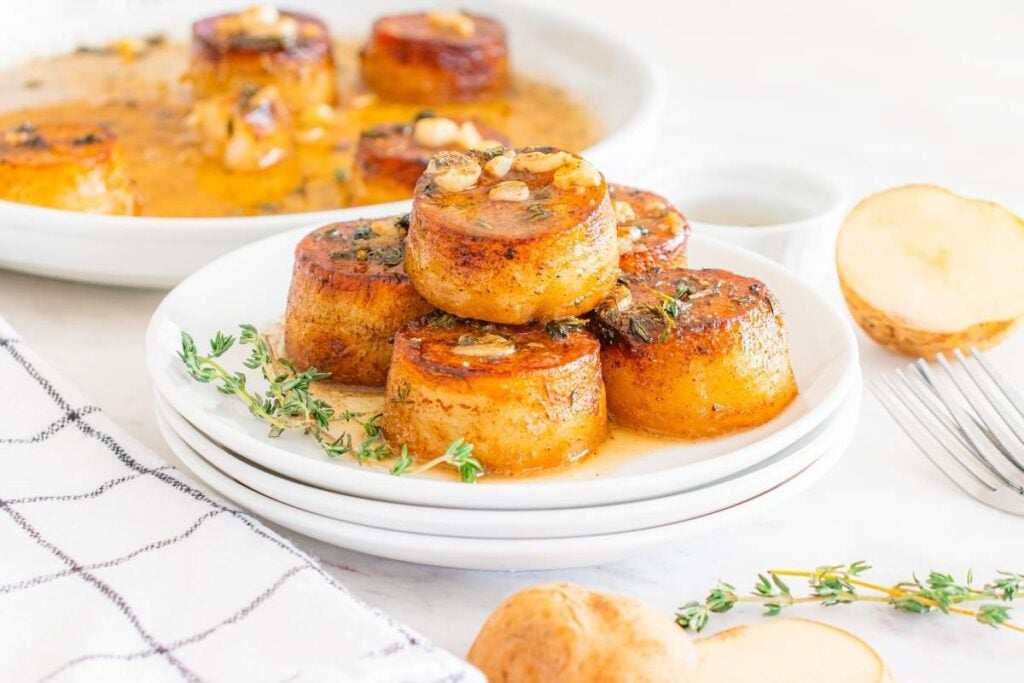 Slices of browned potatoes stacked on a white plate, garnished with herbs and sauce, with more potatoes and a fork nearby.