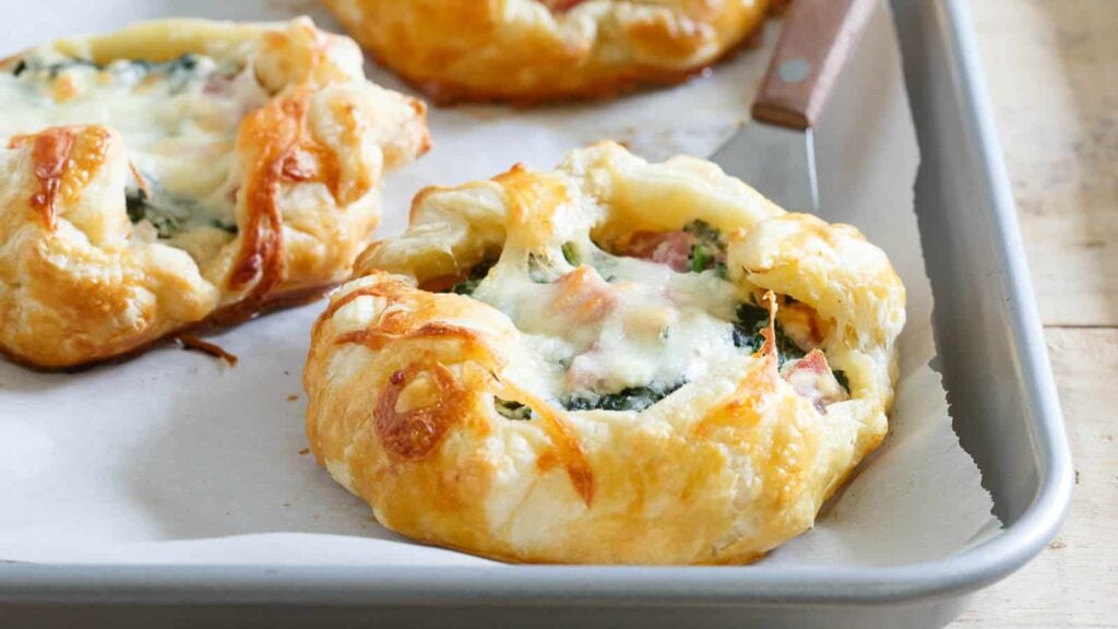 A close-up of a baked pastry filled with spinach, ham, and melted cheese on a baking tray lined with parchment paper.