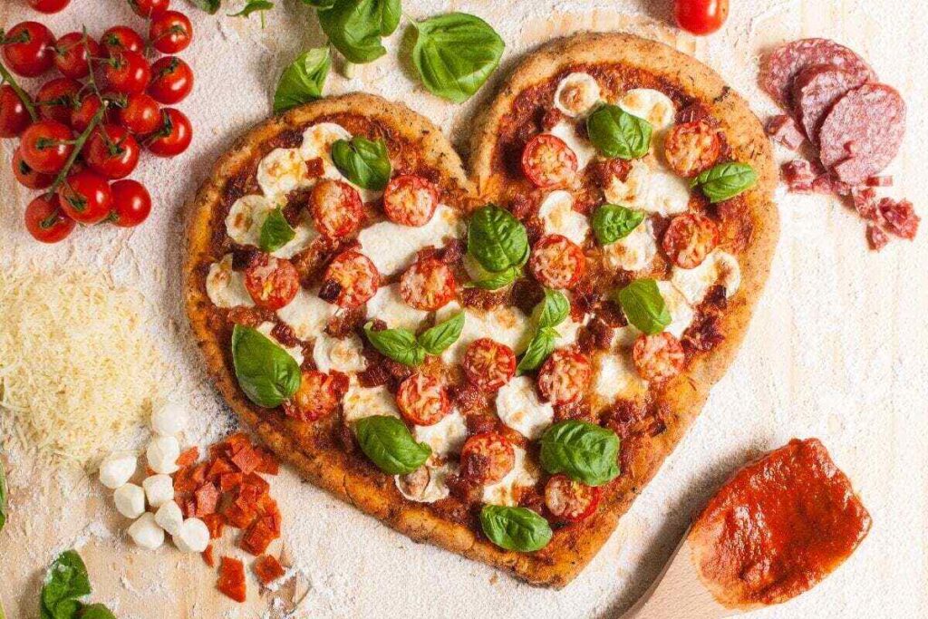 A heart-shaped pizza topped with cherry tomatoes, mozzarella, and basil is surrounded by ingredients like cheese, salami, fresh basil, tomatoes, and marinara sauce on a floured surface.