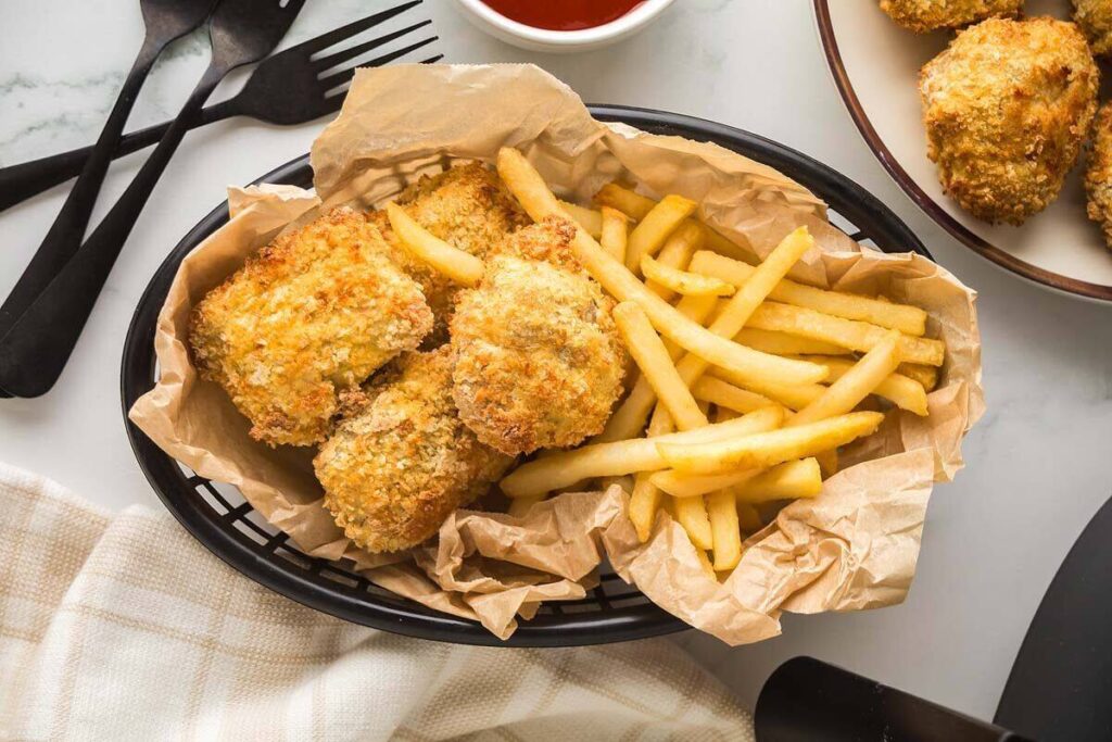 A basket lined with parchment paper holds several pieces of breaded fried chicken and a portion of French fries. Black utensils and a bowl of dipping sauce are nearby.