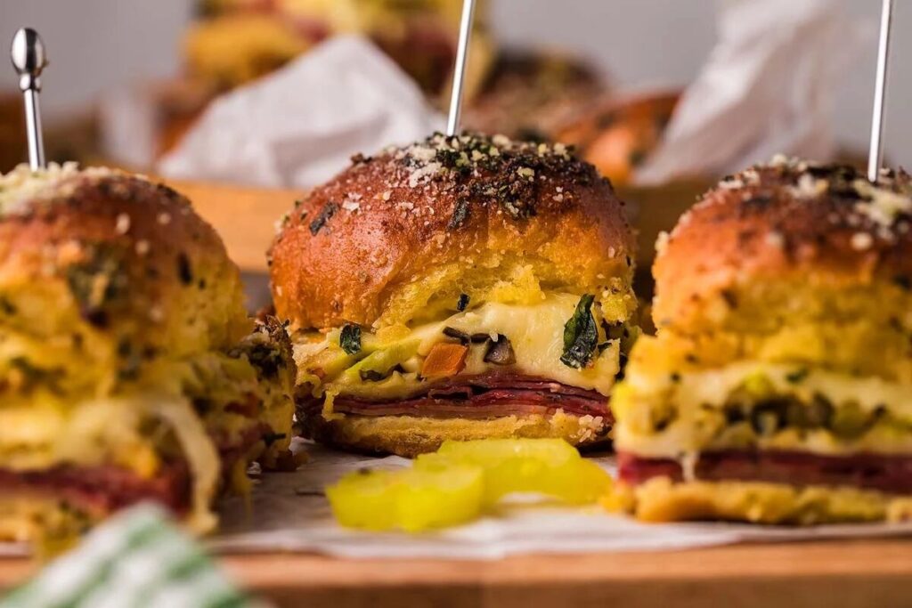 Three small sandwiches with layers of deli meat, cheese, and vegetables on golden buns, topped with herbs and grated cheese, are displayed on a wooden board.