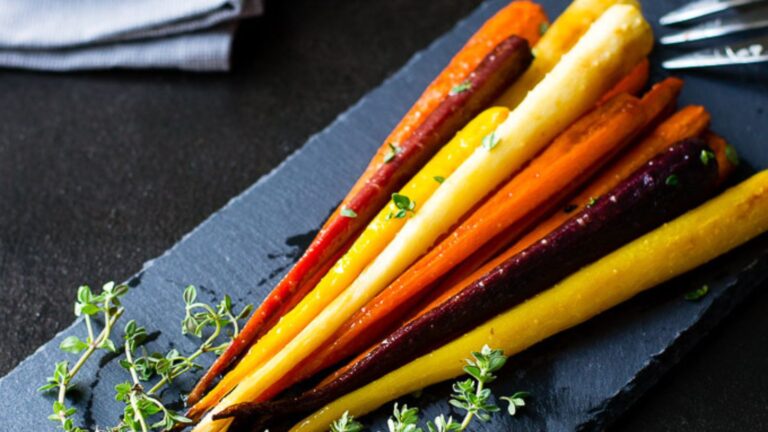A variety of roasted carrots in purple, yellow, and orange colors arranged on a dark slate serving board, garnished with fresh herbs.