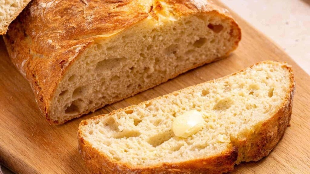 A loaf of rustic bread on a wooden board with a sliced piece in front, topped with a small pat of melting butter.