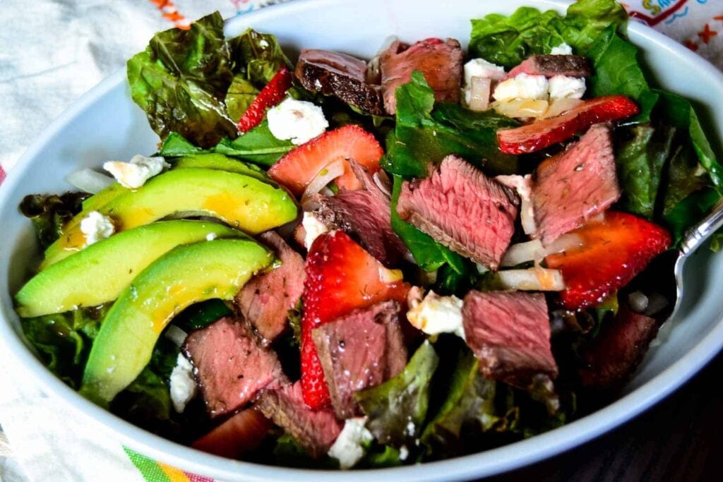 A salad in a white bowl with sliced steak, avocado, strawberries, lettuce, onions, and crumbled cheese.