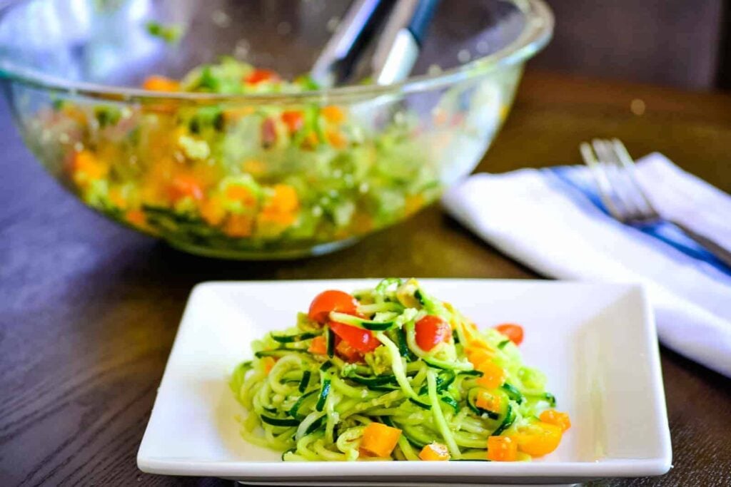 A square white plate with a serving of zucchini noodle salad topped with cherry tomatoes and yellow bell peppers; a glass bowl with more salad is in the background.