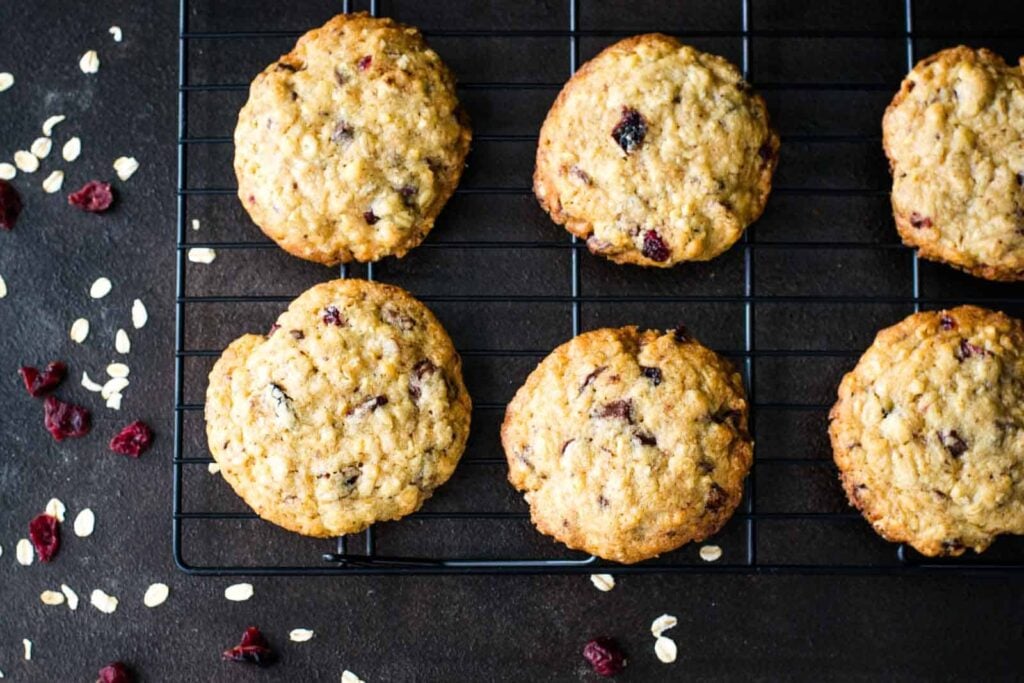 Six oatmeal cookies with visible dried cranberries cooling on a black wire rack, with scattered oats and cranberries on a dark surface.