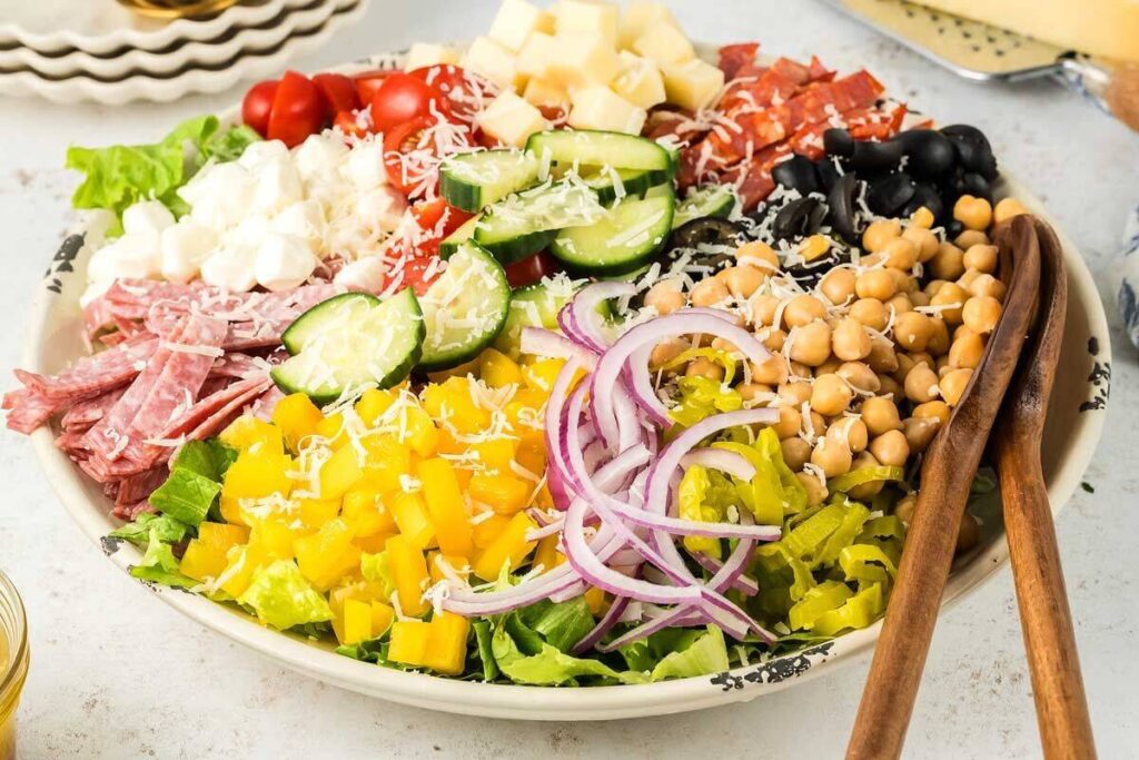 A salad bowl with lettuce, yellow bell peppers, red onions, chickpeas, sliced cucumbers, cherry tomatoes, black olives, cubed cheese, salami, pepperoni, and shredded cheese.