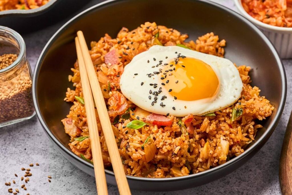 A bowl of kimchi fried rice topped with a sunny-side-up egg and black sesame seeds, served with chopsticks.