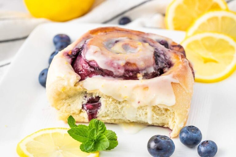 A partially eaten sweet roll with icing, blueberries, and lemon slices on a white plate, garnished with a mint sprig.