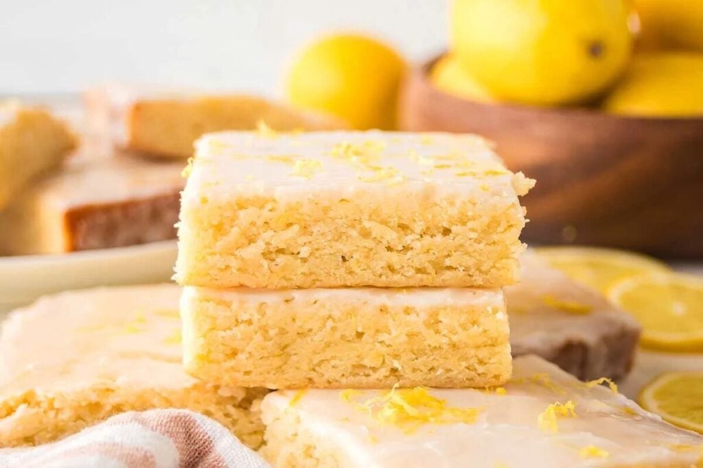 Two lemon bars with icing and lemon zest stacked on top of each other, with more lemon bars and whole lemons in the background.