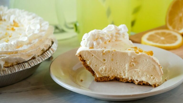 Slice of lemonade pie on a plate with lemonade in the background.