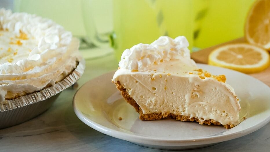 Slice of lemonade pie on a plate with lemonade in the background.