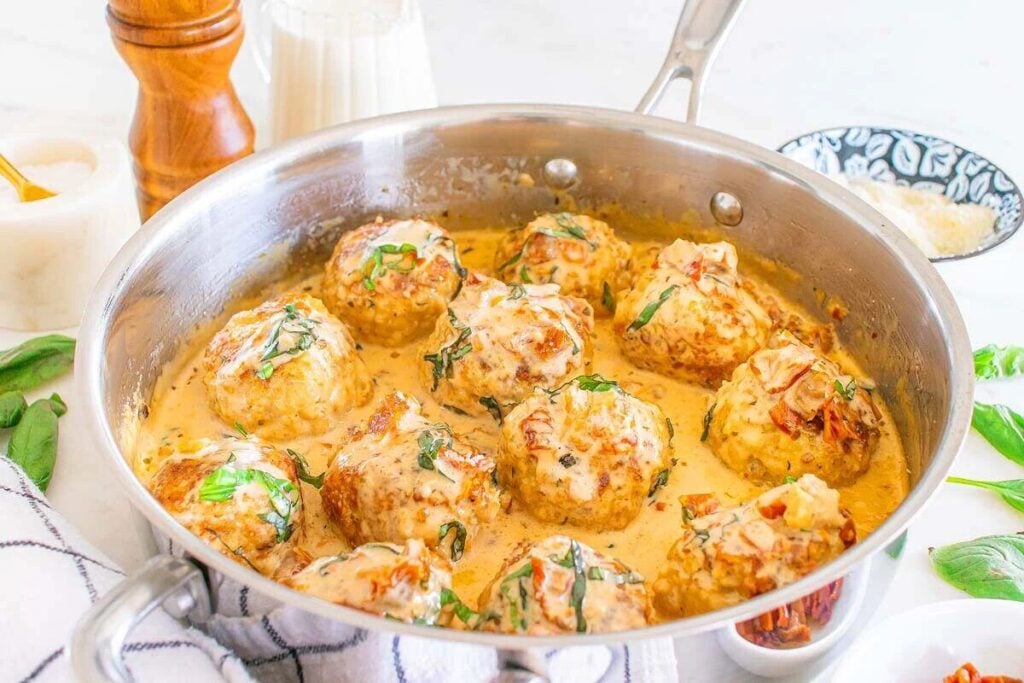 A stainless steel skillet filled with meatballs in a creamy sauce, garnished with chopped herbs, sits on a white surface with various kitchen items nearby.