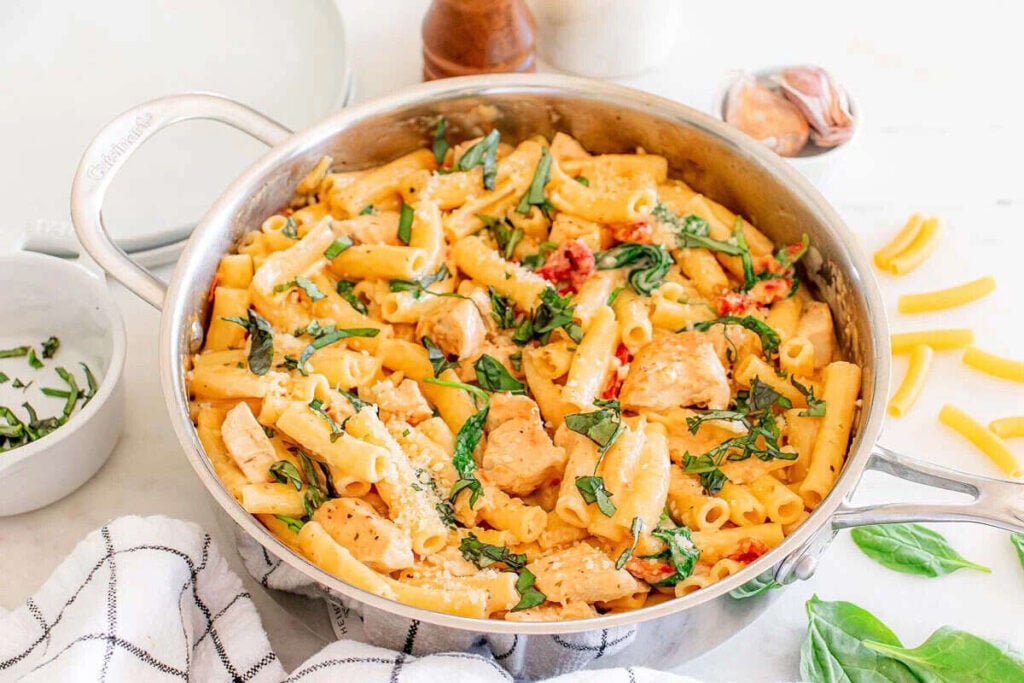 A stainless steel pan filled with rigatoni pasta, cooked chicken pieces, spinach, and a creamy sauce, with fresh basil sprinkled on top.