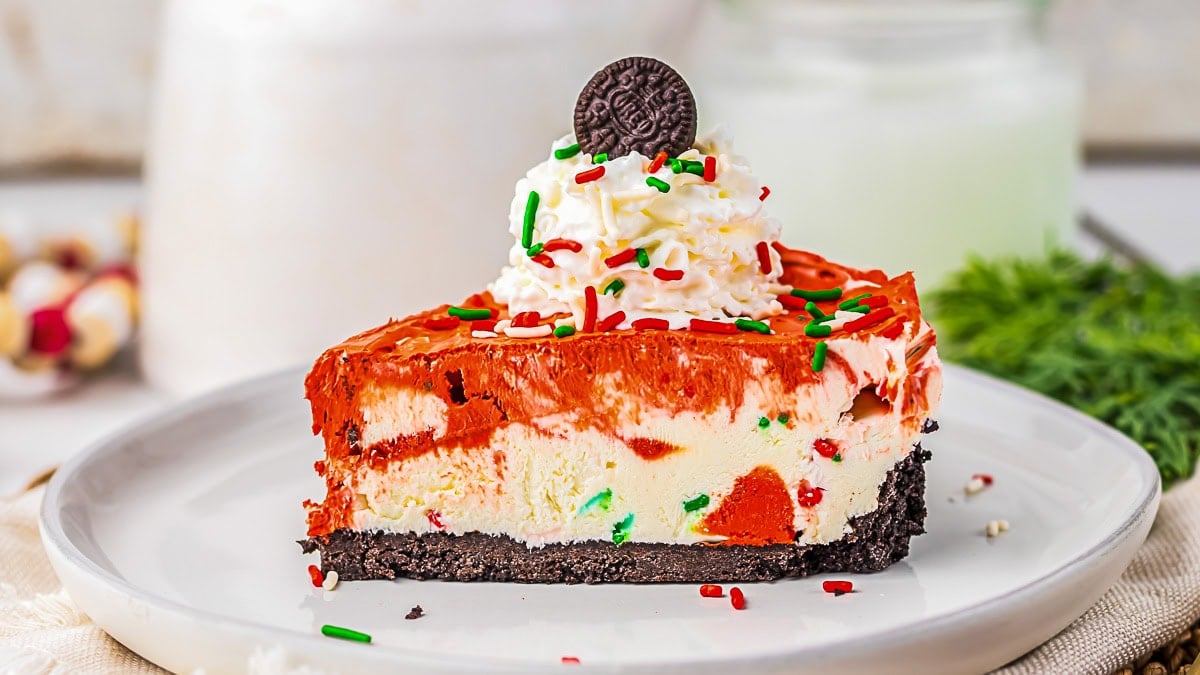 A slice of layered cheesecake with a chocolate cookie crust, red and white filling, whipped cream, colorful sprinkles, and a mini chocolate cookie on top, served on a white plate.