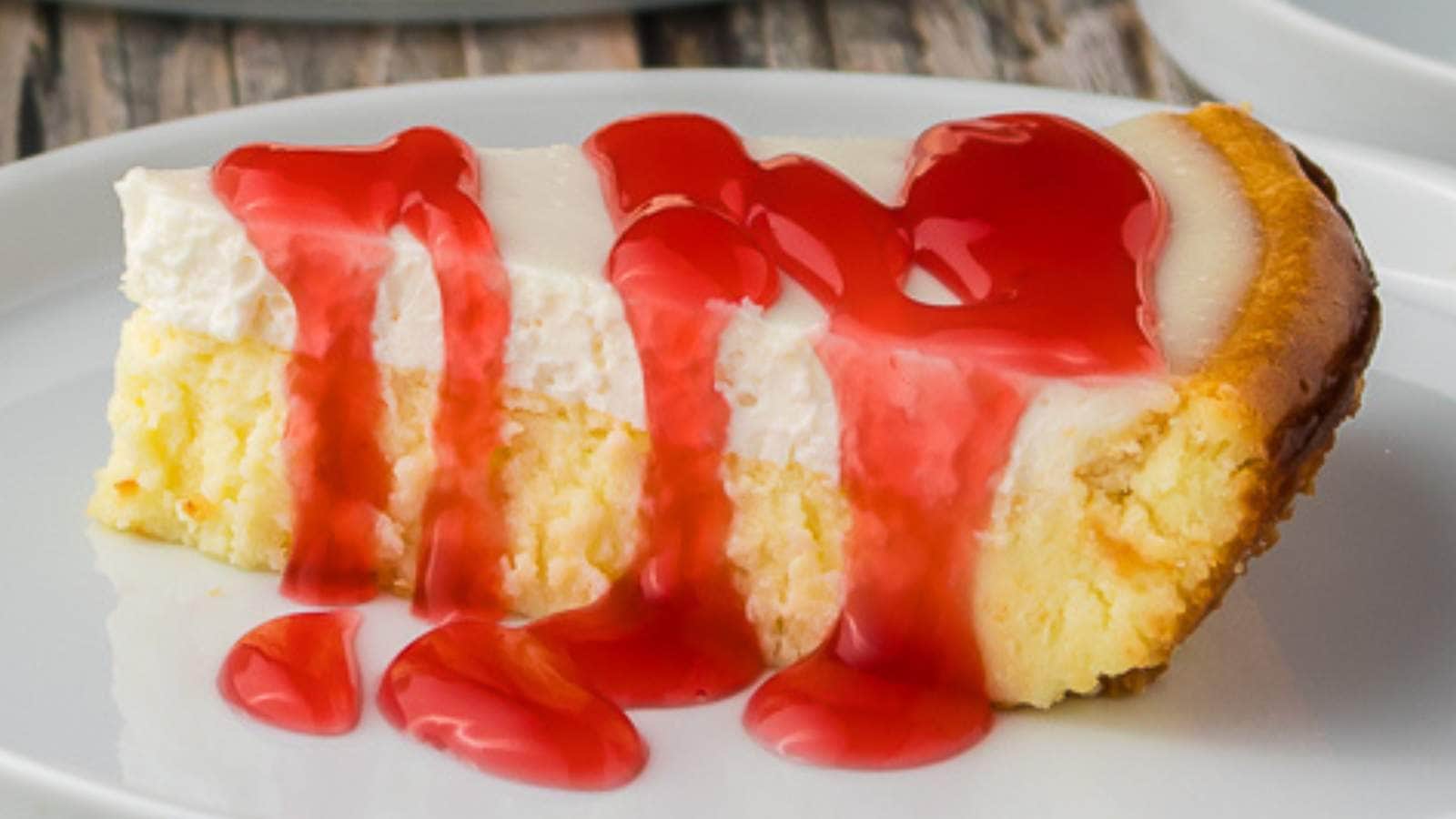 A slice of cheesecake topped with a layer of red strawberry sauce on a white plate.