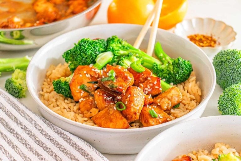 A bowl of brown rice topped with cooked chicken pieces, broccoli florets, sesame seeds, and chopped green onions, with chopsticks resting on the rim.