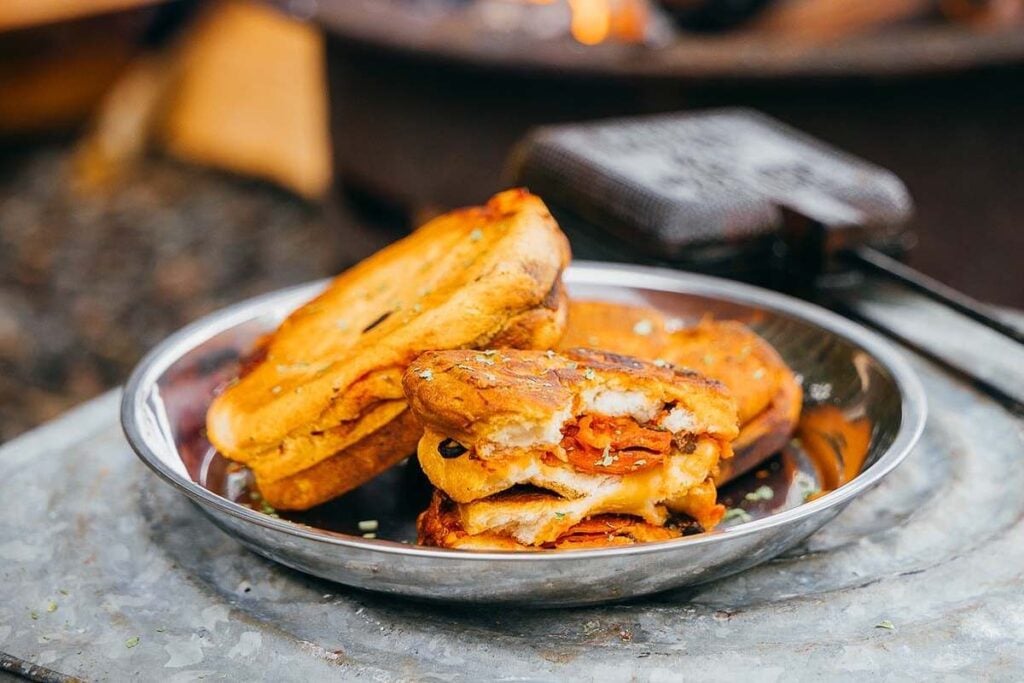 A metal plate with three toasted sandwiches, one partially eaten, showing melted cheese and pepperoni inside, set outdoors.